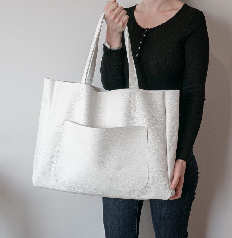 May include: A large, white leather tote bag with a front pocket. The bag has two long handles and is held by a person. The tote bag is a simple, minimalist design, suitable for everyday use. The bag's dimensions are approximately 16 inches wide and 14 inches tall.