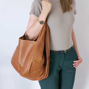 K&ouml;nnte beinhalten: Eine gro&szlig;e, weiche Hobo-Schultertasche aus lohfarbenem Leder mit einer deutlich sichtbaren genarbten Textur. Die Tasche verf&uuml;gt &uuml;ber einen breiten, integrierten Schulterriemen und ein ger&auml;umiges, unstrukturiertes Hauptfach mit weicher Form.