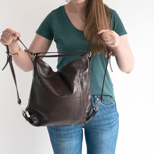 May include: A dark brown leather convertible bag featuring a slouchy, unstructured silhouette with thin adjustable shoulder straps and silver-toned metal hardware. The bag has a smooth finish and a vertical zippered pocket on the front exterior.