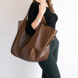 May include: A large, unstructured brown leather tote bag with a smooth finish and wide shoulder straps. The bag features a spacious main compartment and visible exterior stitching, providing a simple and functional design for carrying items.