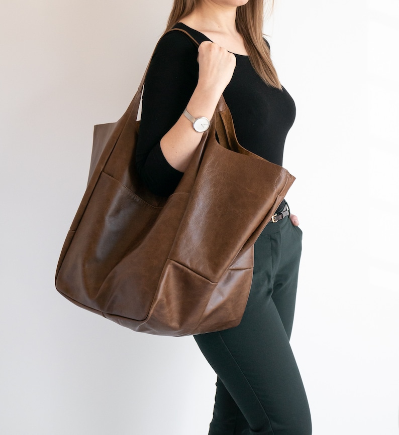 May include: A large, unstructured brown leather tote bag with a smooth finish and wide shoulder straps. The bag features a spacious main compartment and visible exterior stitching, providing a simple and functional design for carrying items.