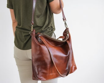 May include: A large, cognac-brown leather hobo bag featuring a textured surface, a top zipper closure, two short carry handles, and a long, adjustable shoulder strap attached with antique brass-toned metal hardware clips.
