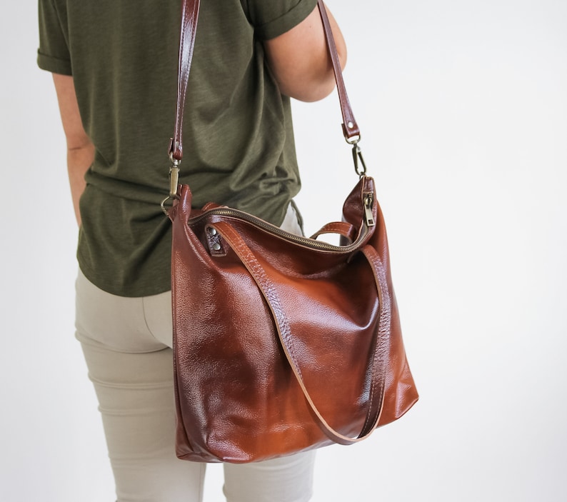 May include: A large, cognac-brown leather hobo bag featuring a textured surface, a top zipper closure, two short carry handles, and a long, adjustable shoulder strap attached with antique brass-toned metal hardware clips.