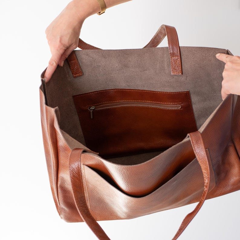 May include: A brown leather tote bag with a zippered pocket on the inside. The bag has two long handles.
