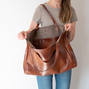 May include: A large brown leather tote bag with a zippered pocket on the inside. The bag is open and the person holding it is showing the inside of the bag.