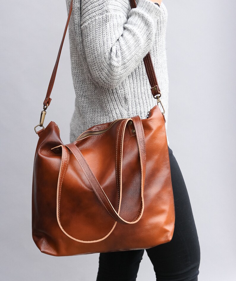 May include: A large, cognac-brown leather tote bag featuring a smooth, polished finish, a top zipper closure, and two sturdy shoulder straps. The bag includes a detachable, adjustable long crossbody strap with antique brass-toned metal hardware.