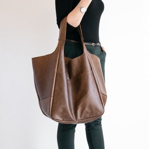 May include: A large, unstructured brown leather tote bag with two wide shoulder straps. The bag features a smooth, matte finish and a spacious, slouchy silhouette with visible seam detailing along the front and sides.