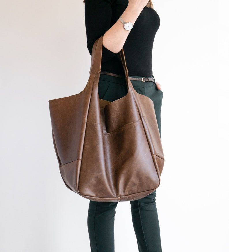 May include: A large, unstructured brown leather tote bag with two wide shoulder straps. The bag features a smooth, matte finish and a spacious, slouchy silhouette with visible seam detailing along the front and sides.