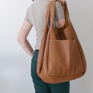 K&ouml;nnte beinhalten: Eine gro&szlig;e, weiche Hobo-Tasche aus hellbraunem Leder mit zwei Schulterriemen und einer auff&auml;lligen Au&szlig;entasche. Die Tasche hat eine genarbte Textur und ein ger&auml;umiges, formloses Design, das locker &uuml;ber der Schulter h&auml;ngt.