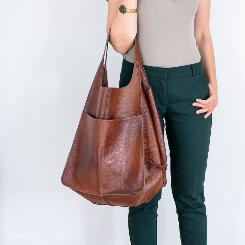 May include: A large brown leather tote bag with a single pocket on the front. The bag is being held by a person wearing a brown shirt and green pants.