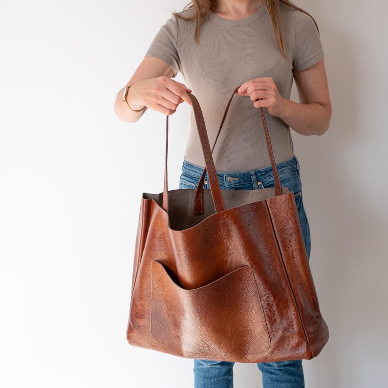 May include: A large brown leather tote bag with a single pocket on the front. The bag has two long straps and is being held by a person.