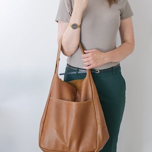 K&ouml;nnte beinhalten: Eine gro&szlig;e, weiche Hobo-Tasche aus braunem Leder mit glatter, genarbter Textur und einem breiten Schulterriemen. Die Tasche verf&uuml;gt &uuml;ber ein tiefes, offenes Hauptfach und eine gro&szlig;e, geschwungene Au&szlig;entasche auf der Vorderseite mit naturfarbenem Innenfutter.