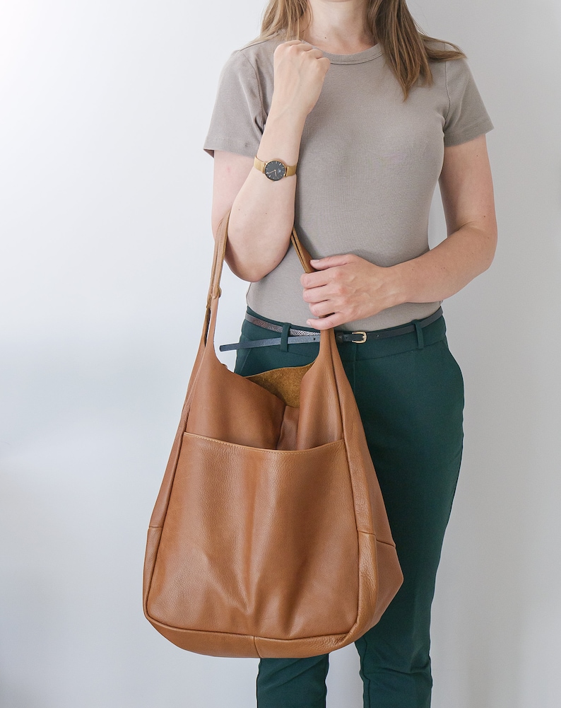 K&ouml;nnte beinhalten: Eine gro&szlig;e, weiche Hobo-Tasche aus braunem Leder mit glatter, genarbter Textur und einem breiten Schulterriemen. Die Tasche verf&uuml;gt &uuml;ber ein tiefes, offenes Hauptfach und eine gro&szlig;e, geschwungene Au&szlig;entasche auf der Vorderseite mit naturfarbenem Innenfutter.