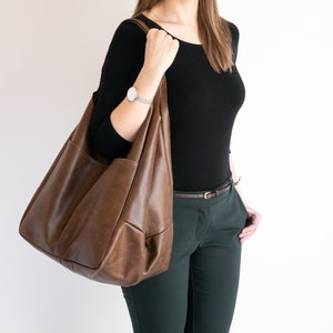 K&ouml;nnte beinhalten: Eine braune Ledertasche mit gro&szlig;em, offenem Oberteil und einem einzigen Trageriemen. Die Tasche wird von einer Person getragen, die ein schwarzes Shirt und eine gr&uuml;ne Hose tr&auml;gt.