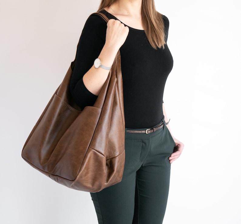 K&ouml;nnte beinhalten: Eine braune Ledertasche mit gro&szlig;em, offenem Oberteil und einem einzigen Trageriemen. Die Tasche wird von einer Person getragen, die ein schwarzes Shirt und eine gr&uuml;ne Hose tr&auml;gt.