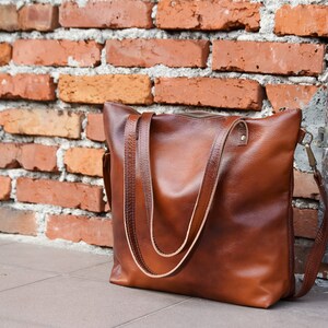 May include: A brown leather tote bag with a smooth texture, featuring two long shoulder straps, a top zipper closure, and a detachable side strap with brass-toned metal hardware attachments on the sides of the bag.