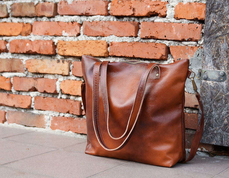 May include: A brown leather tote bag with a smooth texture, featuring two long shoulder straps, a top zipper closure, and a detachable side strap with brass-toned metal hardware attachments on the sides of the bag.