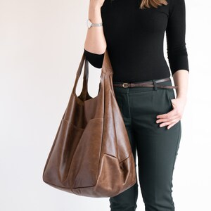 May include: A large, slouchy brown leather hobo bag with a smooth, matte finish and visible stitching details. The bag features a wide shoulder strap and a spacious, unstructured body with a deep, rounded base, designed for carrying everyday items.