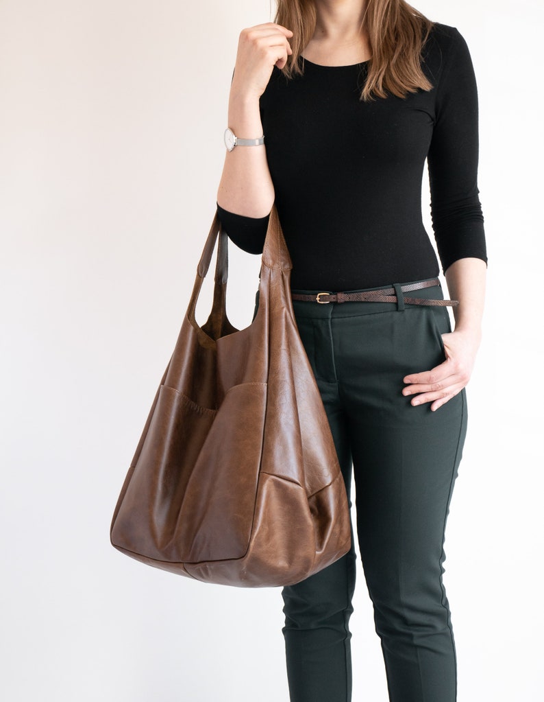 May include: A large, slouchy brown leather hobo bag with a smooth, matte finish and visible stitching details. The bag features a wide shoulder strap and a spacious, unstructured body with a deep, rounded base, designed for carrying everyday items.