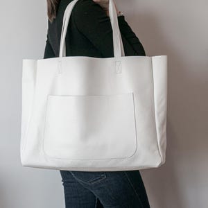 May include: A large, white tote bag with a front pocket and two top handles. The bag is made of a textured material and appears to be empty. The bag is being held by a person wearing a black long-sleeve shirt and blue jeans.