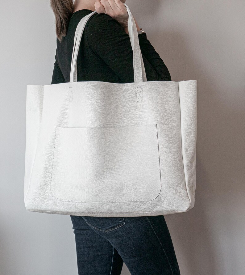 May include: A large, white tote bag with a front pocket and two top handles. The bag is made of a textured material and appears to be empty. The bag is being held by a person wearing a black long-sleeve shirt and blue jeans.