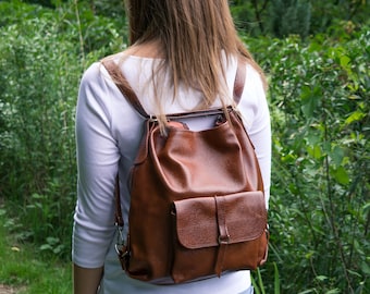 May include: A woman wearing a white shirt and blue jeans is walking in a grassy area. She is carrying a brown leather backpack with a flap closure and a front pocket.