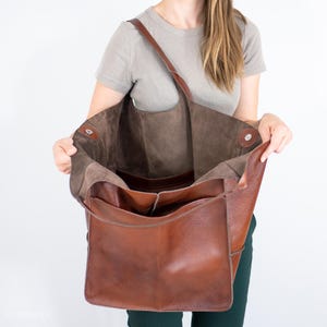 Puede incluir: Un bolso tote grande de cuero marr&oacute;n con forro de ante marr&oacute;n. El bolso est&aacute; abierto y el forro es visible. El bolso tiene una sola correa y un bolsillo con cremallera en la parte delantera.