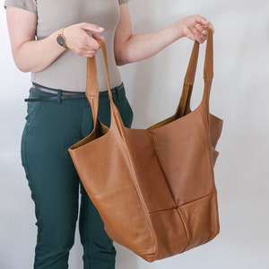 K&ouml;nnte beinhalten: Eine gro&szlig;e, unstrukturierte Tragetasche aus hellbraunem Leder mit zwei langen Schulterriemen. Die Tasche hat eine weiche, genarbte Textur und ein ger&auml;umiges Design mit offener Oberseite und sichtbaren N&auml;hten.