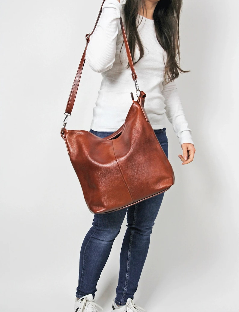 May include: A brown leather tote bag with a long adjustable strap. The bag is worn over the shoulder of a person wearing jeans and a white shirt.