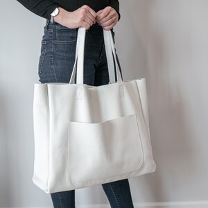 May include: A large, white leather tote bag with a front pocket and two long handles. The bag is held by a person wearing a black long-sleeve shirt and dark blue jeans. The bag is a neutral color and appears to be made of a soft material.