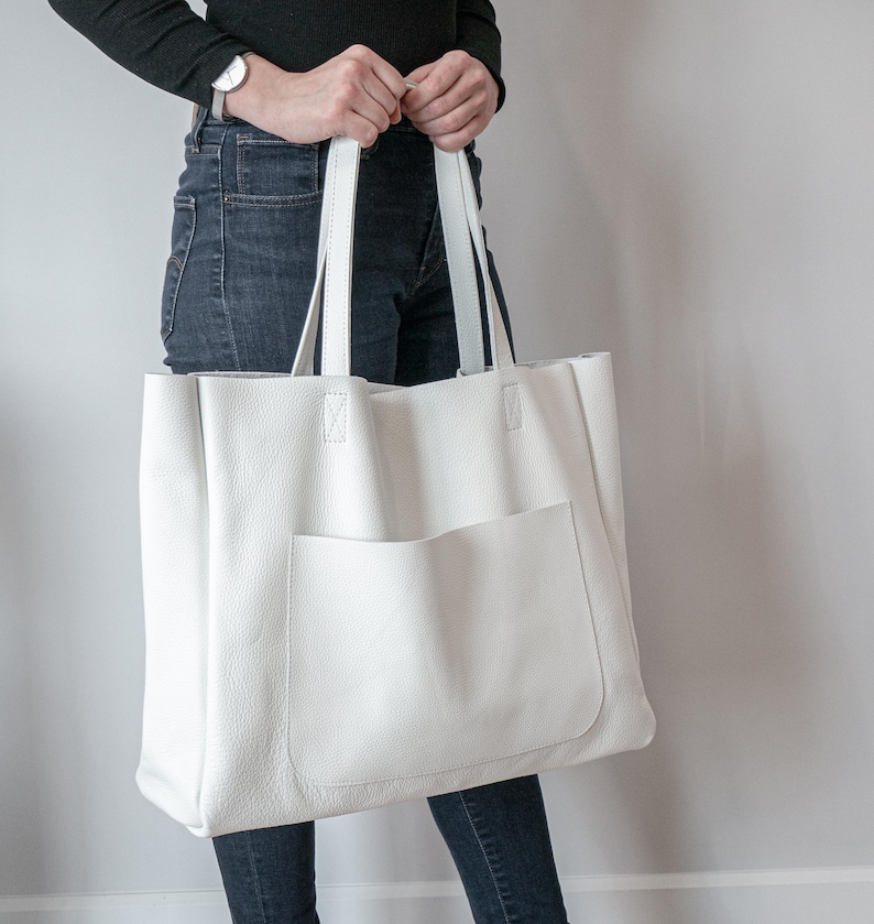 May include: A large, white leather tote bag with a front pocket and two long handles. The bag is held by a person wearing a black long-sleeve shirt and dark blue jeans. The bag is a neutral color and appears to be made of a soft material.