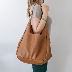 K&ouml;nnte beinhalten: Eine gro&szlig;e, weiche Hobo-Schultertasche aus glattem, lohfarbenem Leder. Die Tasche verf&uuml;gt &uuml;ber einen breiten, integrierten Schulterriemen und eine tiefe, offene Au&szlig;entasche auf der Vorderseite, die eine weiche, unstrukturierte Silhouette aufweist.