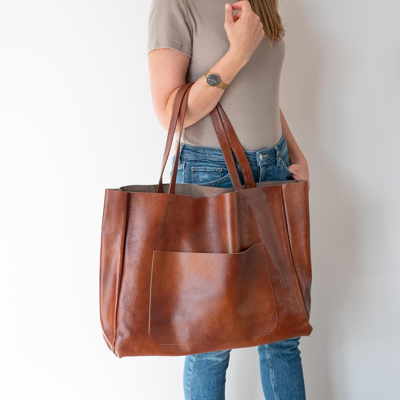 May include: A large brown leather tote bag with a single front pocket. The bag has a relaxed, slouchy look and is made of a high-quality leather.