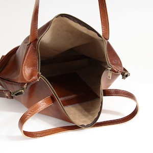 May include: A brown leather tote bag with a zippered top opening, two shoulder straps, and a beige interior lining. The bag features a smooth, textured exterior surface and a single interior pocket visible inside the main compartment.