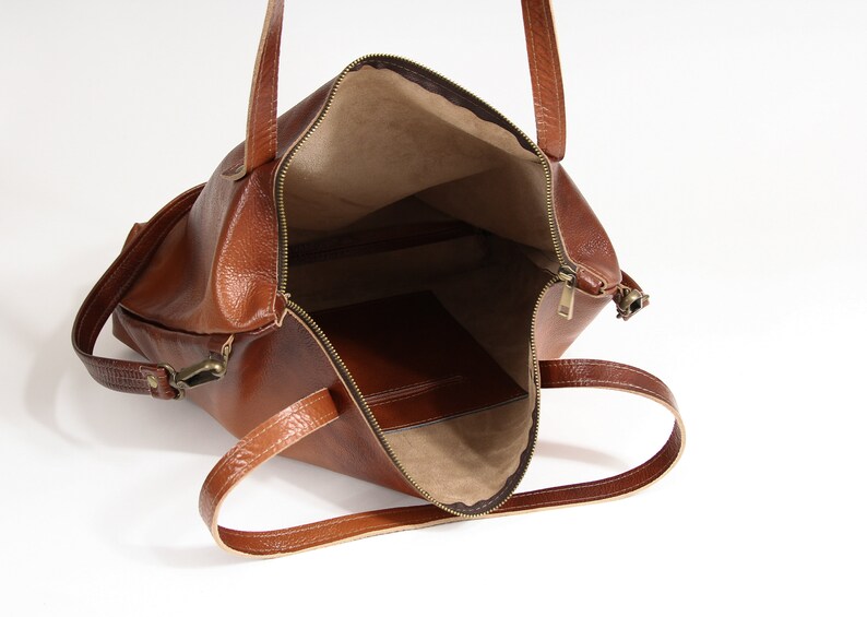 May include: A brown leather tote bag with a zippered top opening, two shoulder straps, and a beige interior lining. The bag features a smooth, textured exterior surface and a single interior pocket visible inside the main compartment.