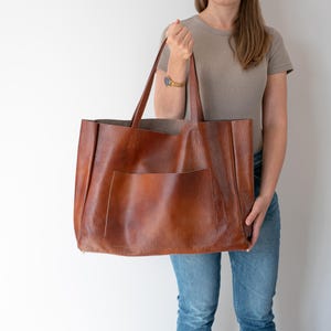 May include: A large brown leather tote bag with a single pocket on the front. The bag has a wide opening and two handles.
