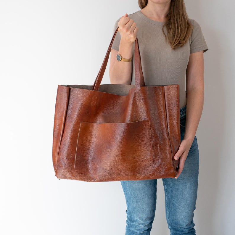 May include: A large brown leather tote bag with a single pocket on the front. The bag has a wide opening and two handles.