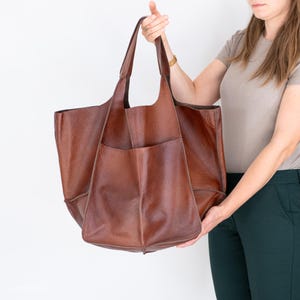 May include: A large brown leather tote bag with a single pocket on the front. The bag has a wide opening and two handles.