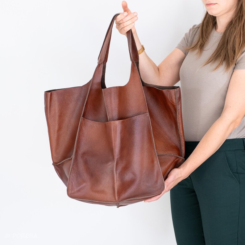 May include: A large brown leather tote bag with a single pocket on the front. The bag has a wide opening and two handles.
