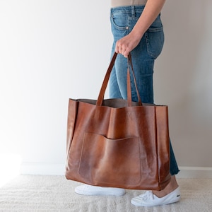 May include: A large brown leather tote bag with a single pocket on the front. The bag has two long handles and is being held by a person wearing blue jeans.