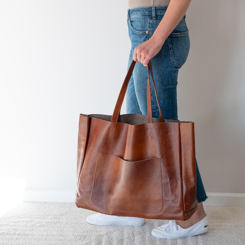 May include: A large brown leather tote bag with a single pocket on the front. The bag has two long handles and is being held by a person wearing blue jeans.