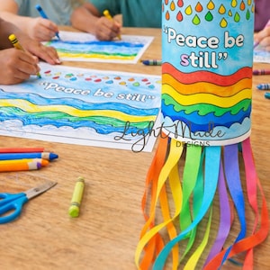 May include: A cylindrical craft project with a blue sky design, rainbow, and the words "Peace be still." Colorful streamers hang from the bottom. Children are coloring similar designs on paper with crayons.