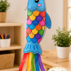 May include: A colorful, handmade kite shaped like a fish, with a blue head and tail, and rainbow-colored scales. The kite has rainbow streamers and the text "Printable Fishers of men kite Craft" and "Digital Download".