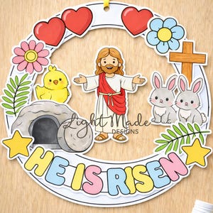 May include: A printable Easter craft with a circular design and the text "He is Risen." The design includes illustrations of Jesus, hearts, flowers, bunnies, a chick, a tomb, and stars. A yellow ribbon is attached for hanging.