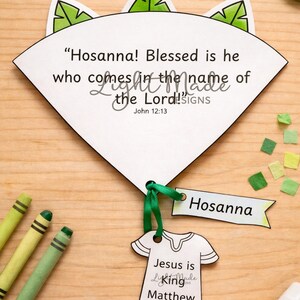 May include: A white paper craft with the text "Hosanna! Blessed is he who comes in the name of the Lord!" and "Jesus is King". Includes a green ribbon, a "Hosanna" tag, and crayons. The craft is on a wooden surface.