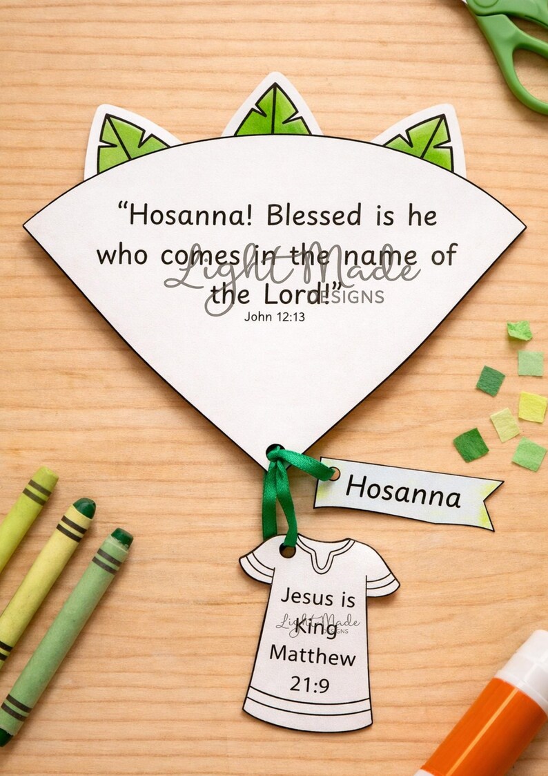 May include: A white paper craft with the text "Hosanna! Blessed is he who comes in the name of the Lord!" and "Jesus is King". Includes a green ribbon, a "Hosanna" tag, and crayons. The craft is on a wooden surface.