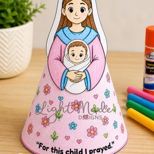 Puede incluir: Una manualidad de papel imprimible con una ilustración de dibujos animados de una mujer sosteniendo a un bebé. La mujer viste un atuendo rosa y azul, y la base en forma de cono está decorada con flores y el texto "For this child I prayed."