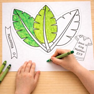 May include: A coloring page with three large leaf outlines, partially colored green. Text on the page includes "Hosanna" and "Jesus is King Matthew 21:9." A child's hand is coloring with a green crayon. Green crayons and scissors are also visible.