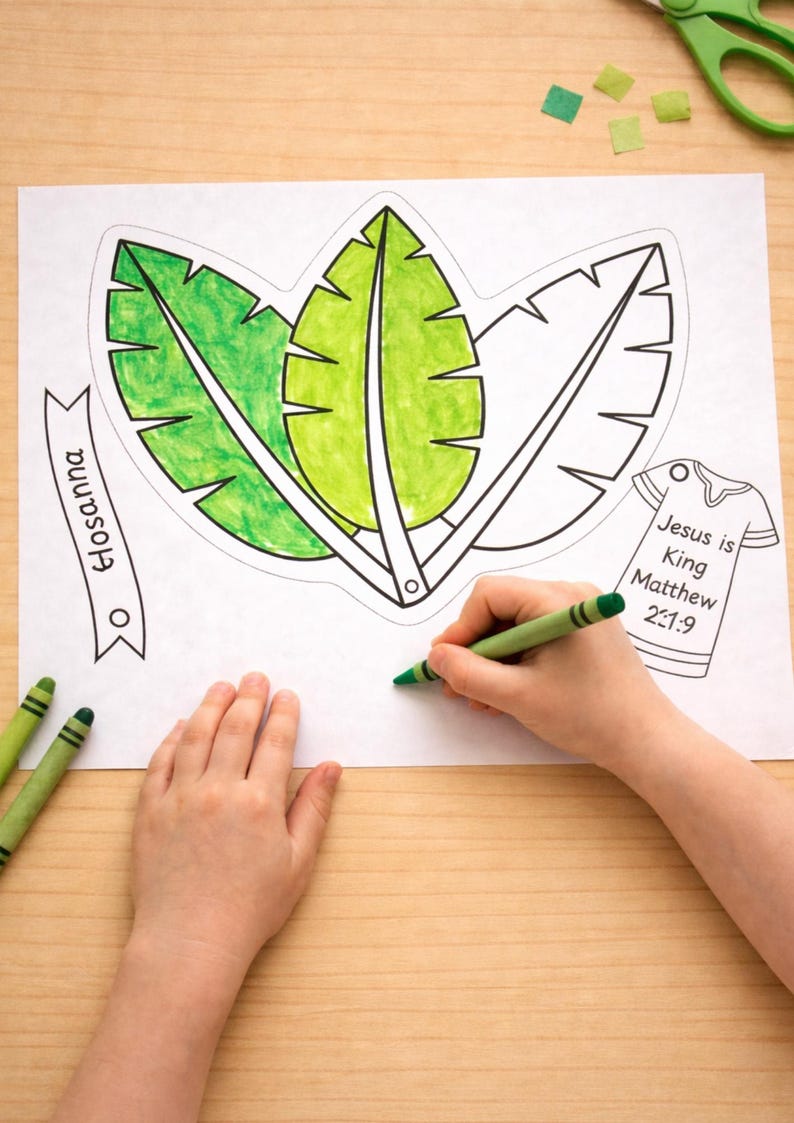 May include: A coloring page with three large leaf outlines, partially colored green. Text on the page includes "Hosanna" and "Jesus is King Matthew 21:9." A child's hand is coloring with a green crayon. Green crayons and scissors are also visible.