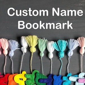 Puede incluir: Una fila de borlas de hilo de colores con letras de fieltro unidas, deletreando "Custom Name Bookmark".